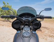 A close-up, eye-level view of the front of a dark gray RWD V-Twin motorcycle, featuring a black fairing with integrated speakers, a clear windshield, black handlebars with mirrors, and chrome gauges on the fuel tank. The motorcycle is parked outdoors on a dirt path with a tree and blue sky in the background.